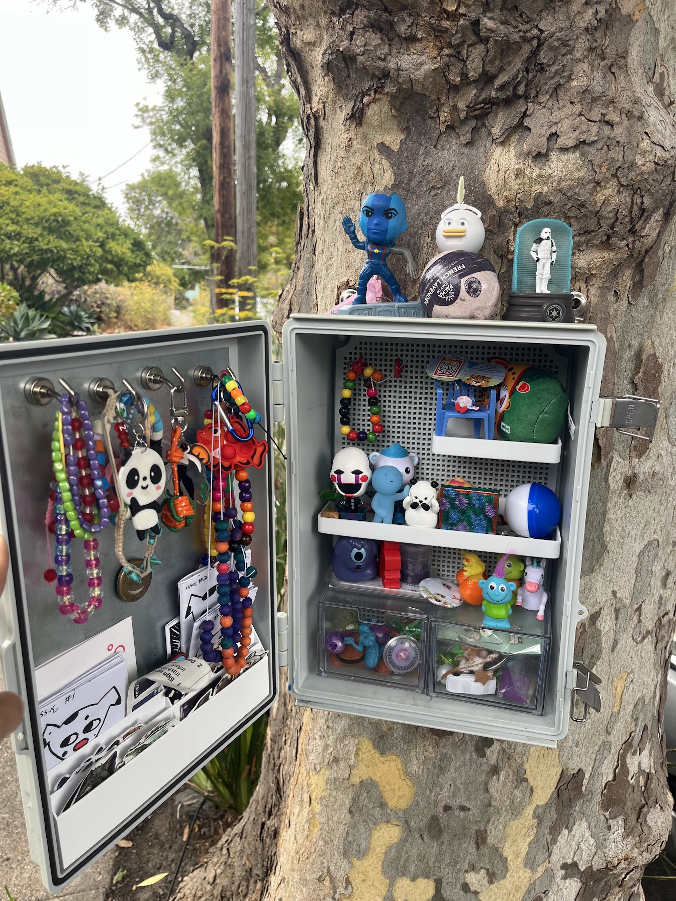 The Berkeley Trinket Trade box mounted on a tree trunk, its hinged door open to show necklaces hung on hooks and small bins of figurines on a grid back panel